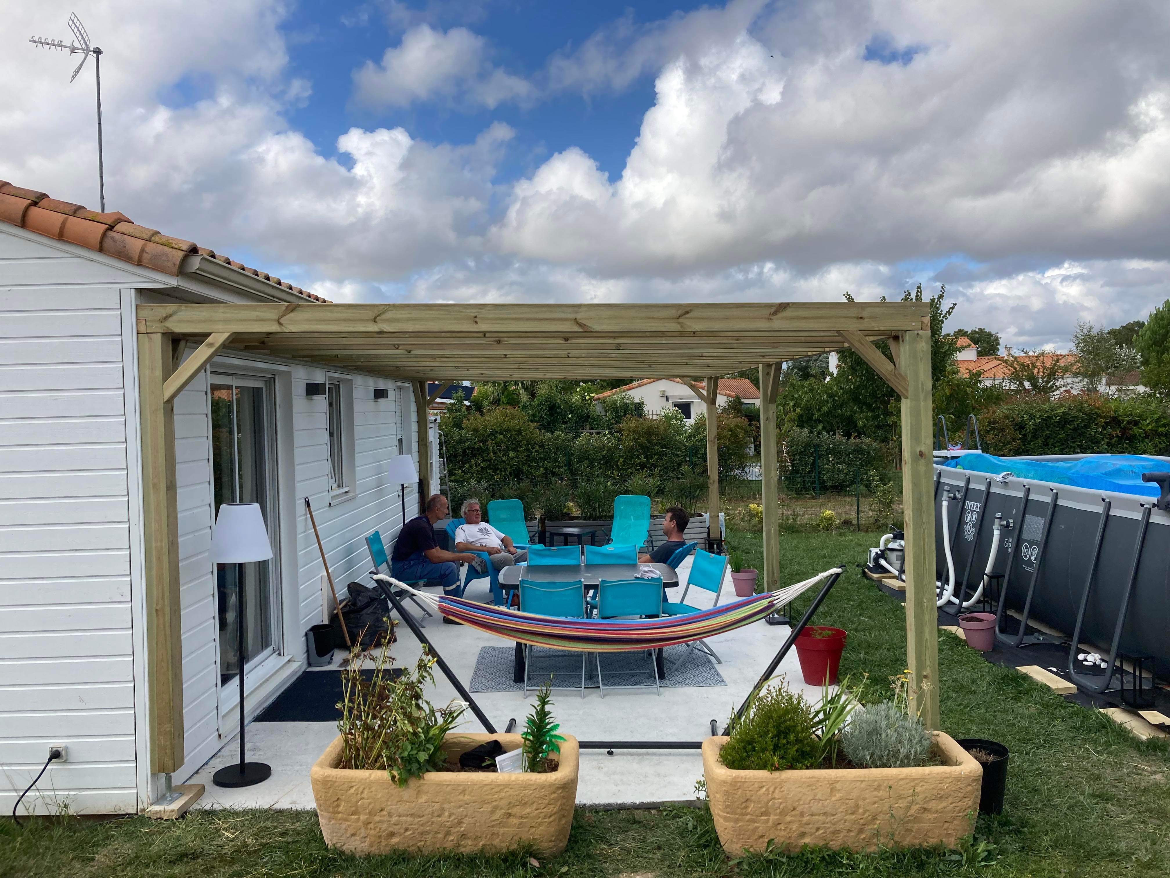 Pergola bois adossée à une maison blanche avec des personnes détendues sur une terrasse